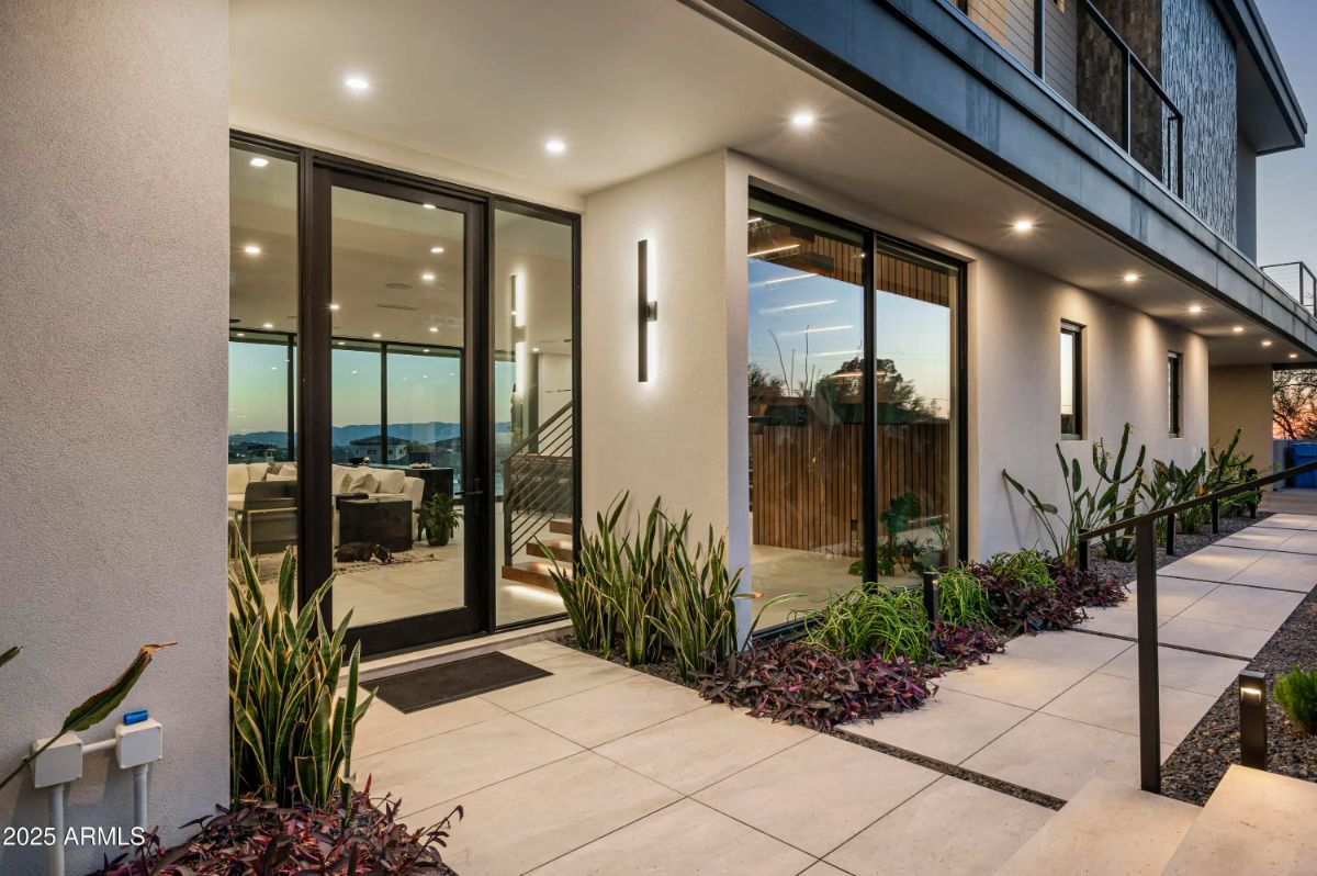 Modern front entry with large glass doors, sleek lighting, and desert landscaping lining a tiled walkway. 