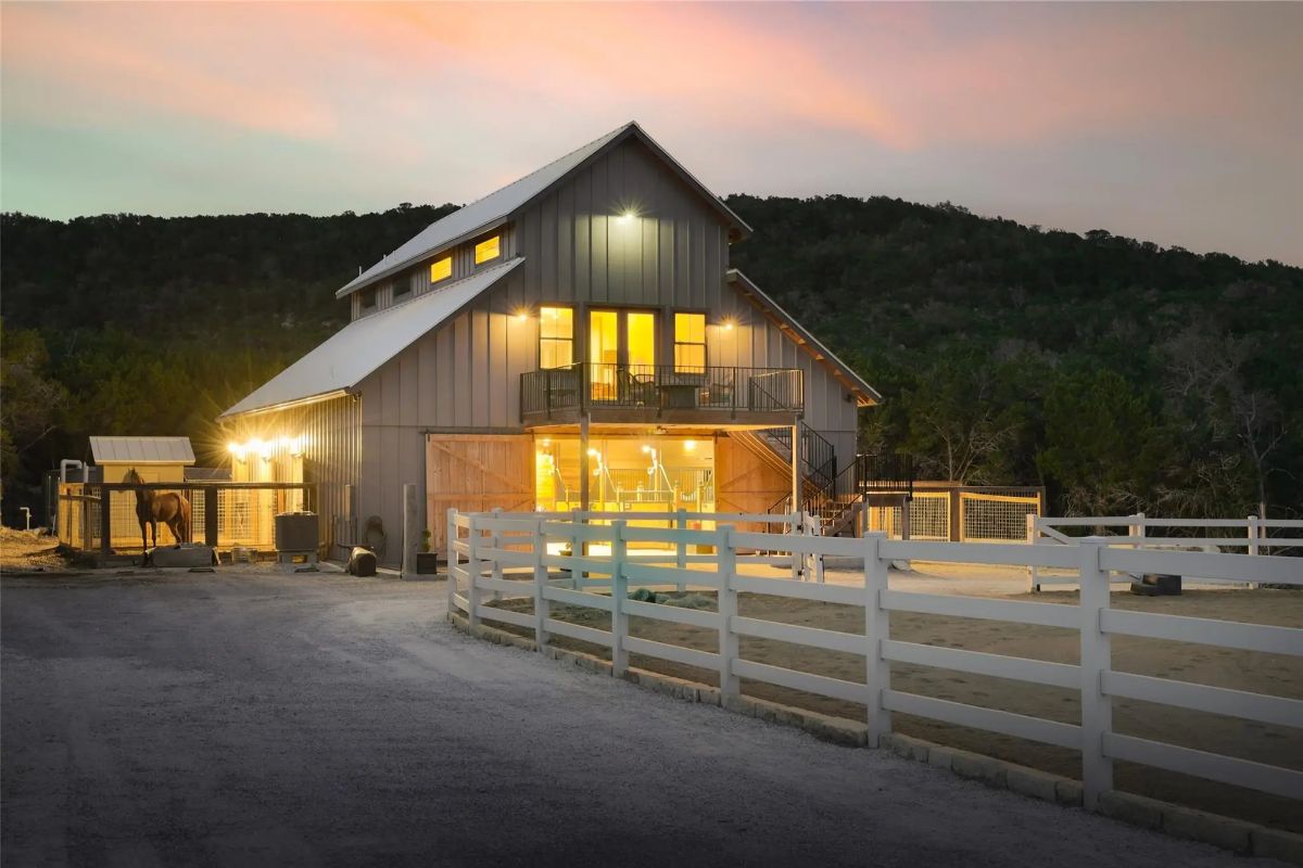 Tall barn with board-and-batten siding sits adjacent to a white fenced arena.
