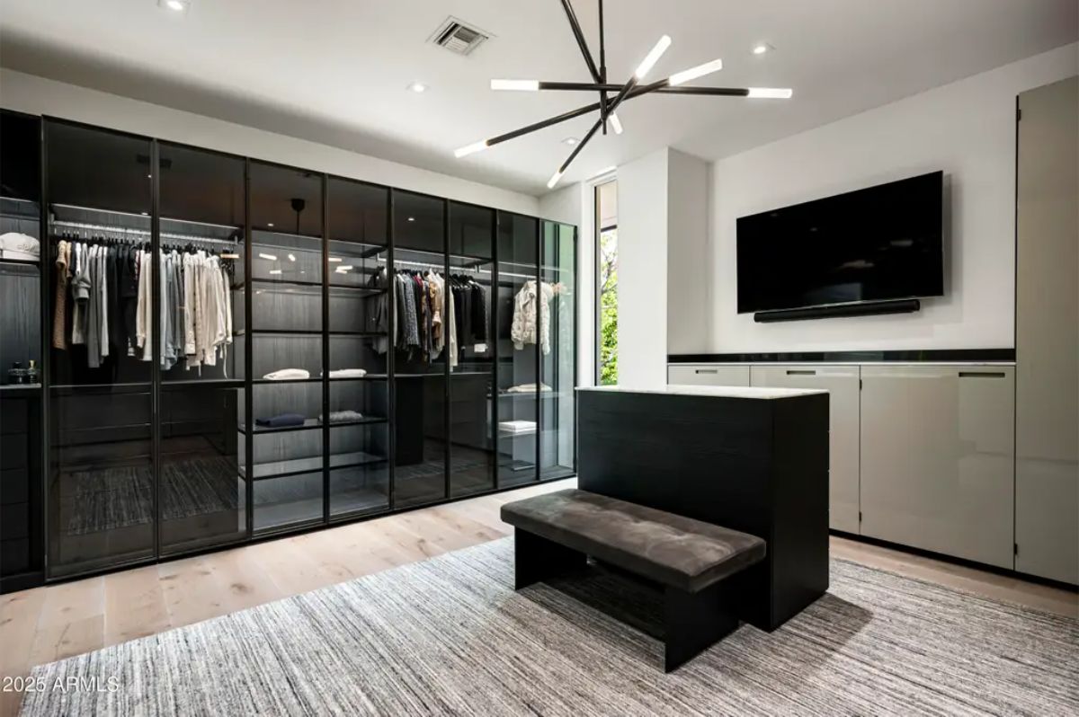 Spacious walk-in closet with glass-front wardrobes and central bench sits beneath modern lighting. 