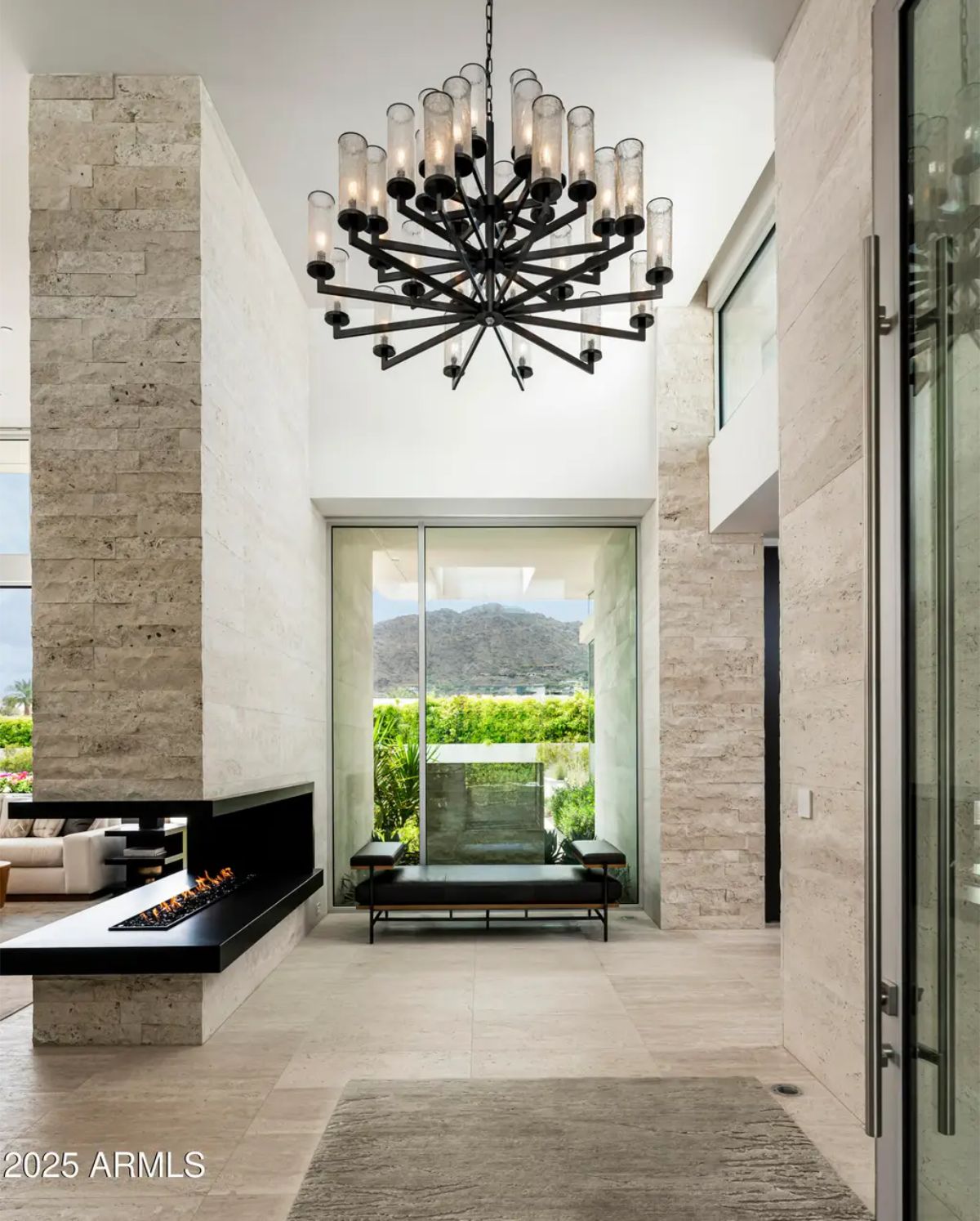 Grand entry with towering ceilings, black sculptural chandelier, stone walls, and mountain views through glass doors. 
