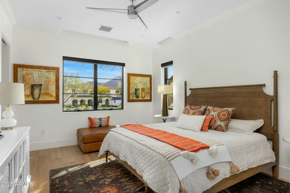 Guest bedroom with ceiling fan, warm accent tones, and large window overlooking landscaped grounds. 