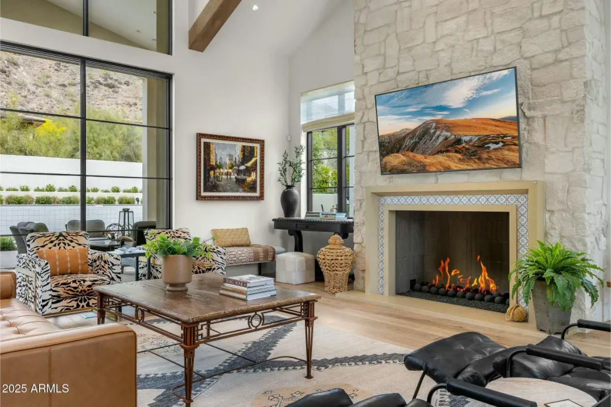 Living room with tall windows, vaulted ceiling, stone fireplace, and a mix of modern and classic furnishings. 