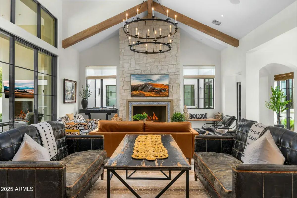 Great room with exposed beams, chandelier, stone fireplace, and twin leather sofas arranged around a central table. 