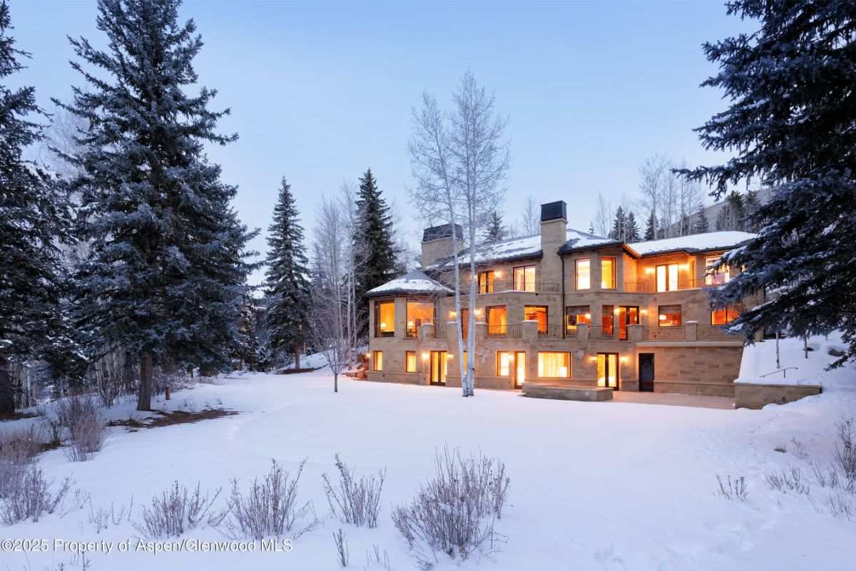 Evening view of snow-covered estate with glowing interior lights nestled among tall trees. 