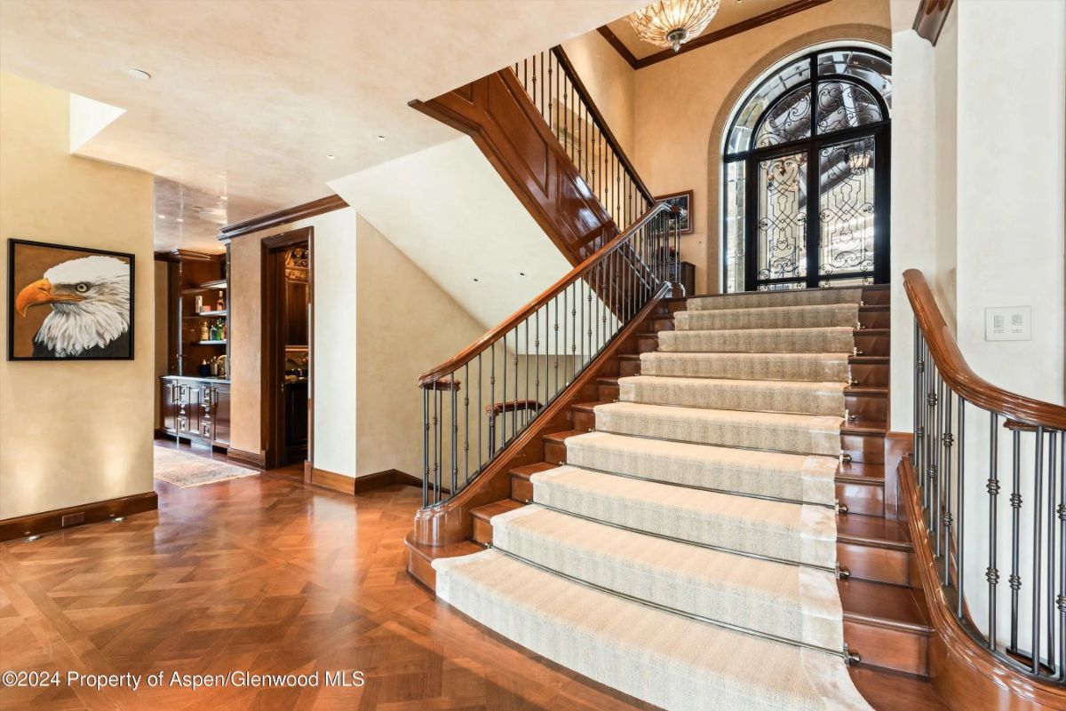 Grand staircase with wood treads, carpet runner, and curved iron railing beneath a chandelier. 
