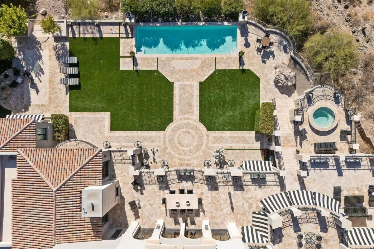 Aerial view of an intricately designed backyard with symmetrical layout, rectangular pool, spa, outdoor kitchen, and striped lounge areas surrounded by stone pavers and artificial turf. 