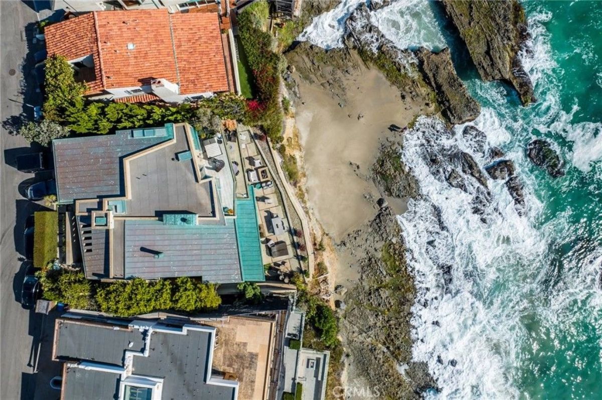 Modern home with angular rooflines sits directly above a sandy beach and rocky shoreline.