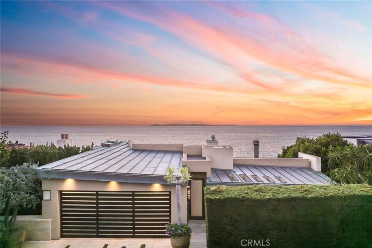 Modern residence with flat metal roof sits behind a hedge and gate.