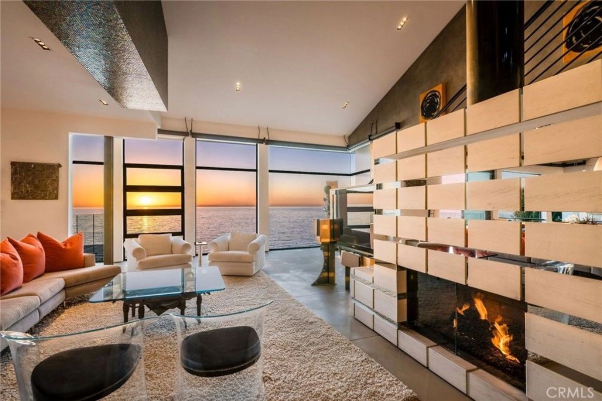 Ocean and sunset are visible through large modern windows behind lounge chairs.