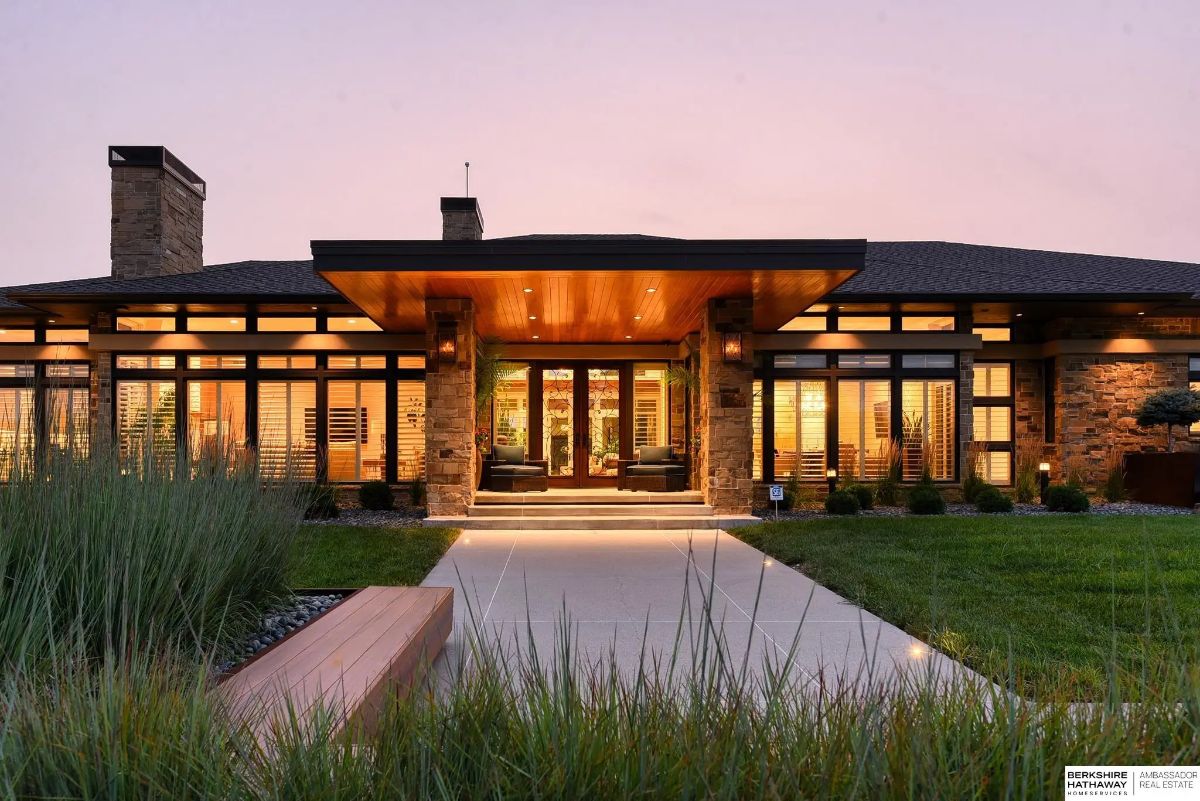 Front exterior view with overhang entry canopy, stone façade, large windows, and landscaped path at dusk.