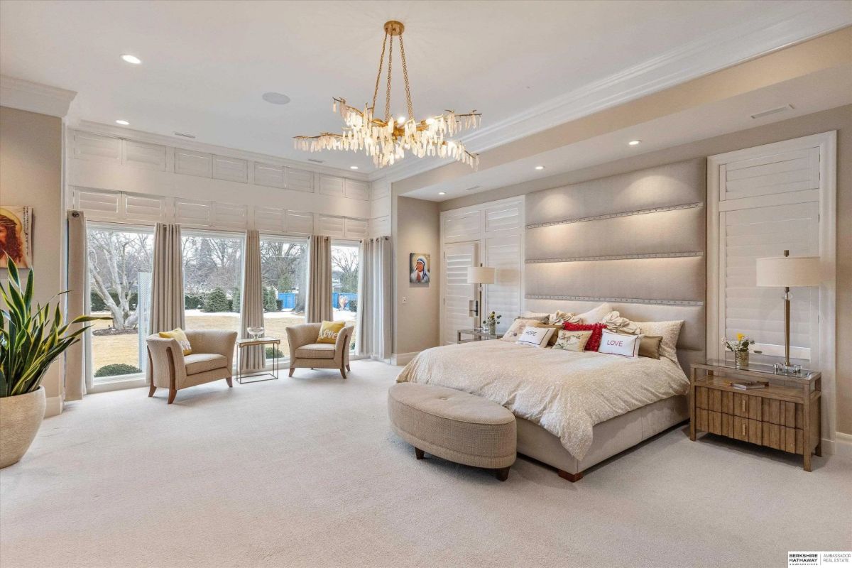 Primary bedroom with upholstered headboard wall, chandelier, seating area, and large windows with shutters.