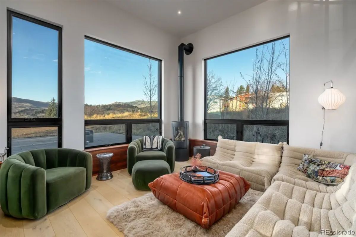 Lounge area features green and beige seating, corner fireplace, and expansive windows with views of nearby homes and hills.