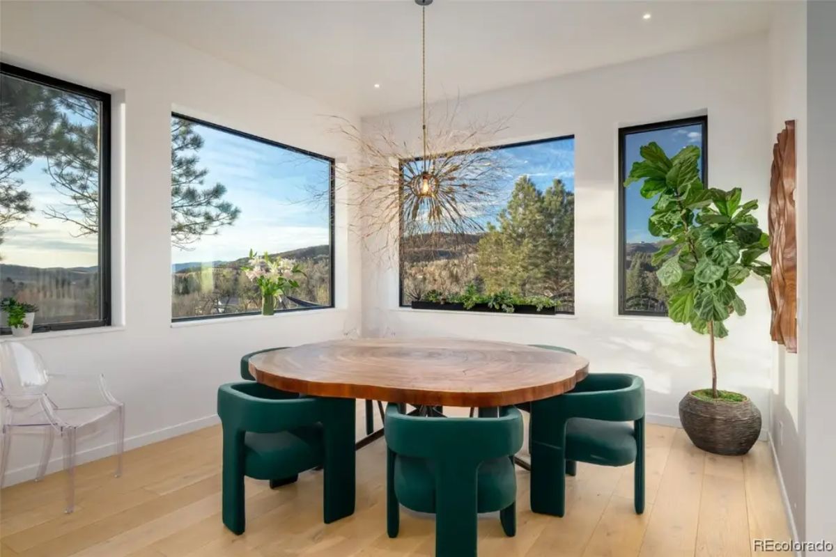Dining area with round wood table and green upholstered chairs is surrounded by large windows and modern chandelier.
