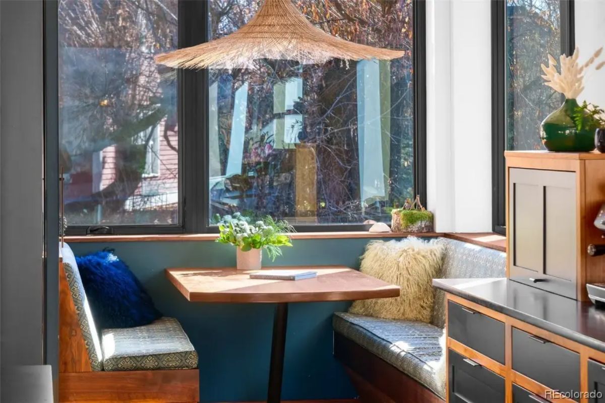 Kitchen nook with built-in banquette seating and wood table is lit by a woven pendant light and surrounded by tall windows.