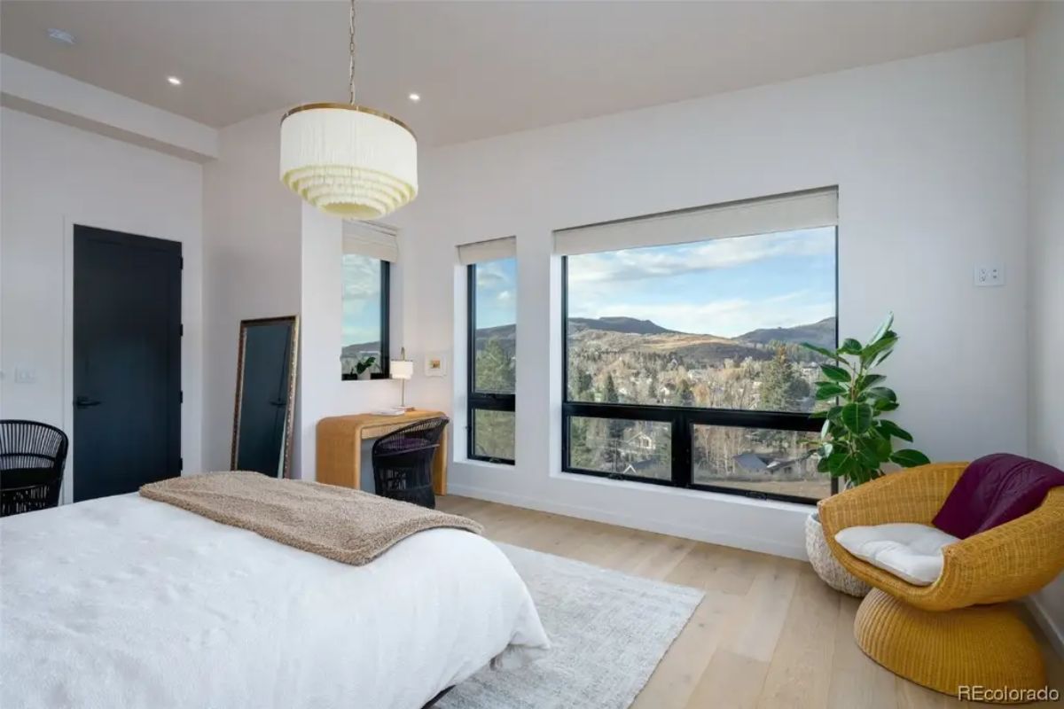 Bedroom features light wood flooring, large window with mountain view, white bed, wicker chair, and modern pendant light.