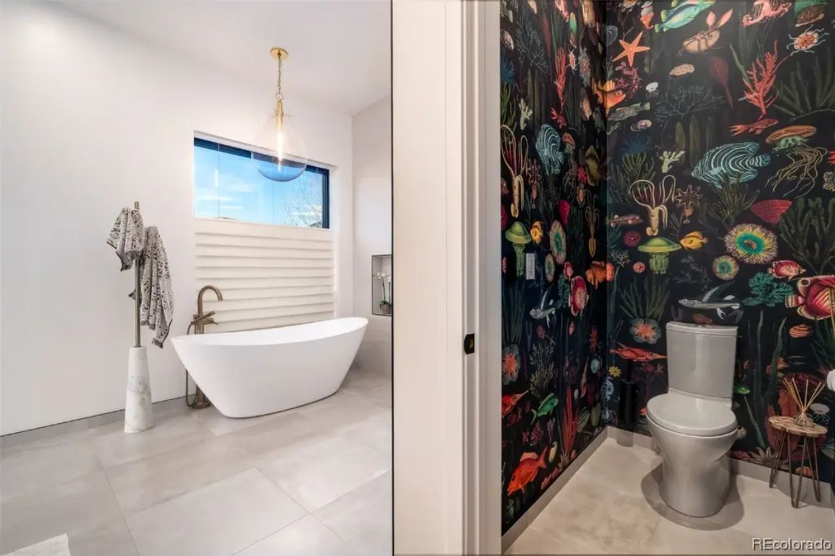 Bathroom with freestanding tub near window on the left and separate toilet room with colorful ocean-themed wallpaper on the right.
