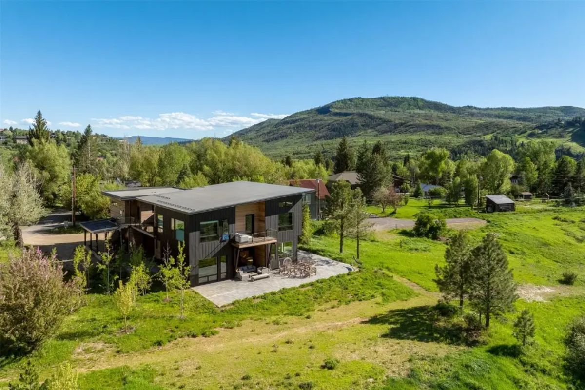 Modern house with angular roof sits on large grassy lot surrounded by trees and hills.