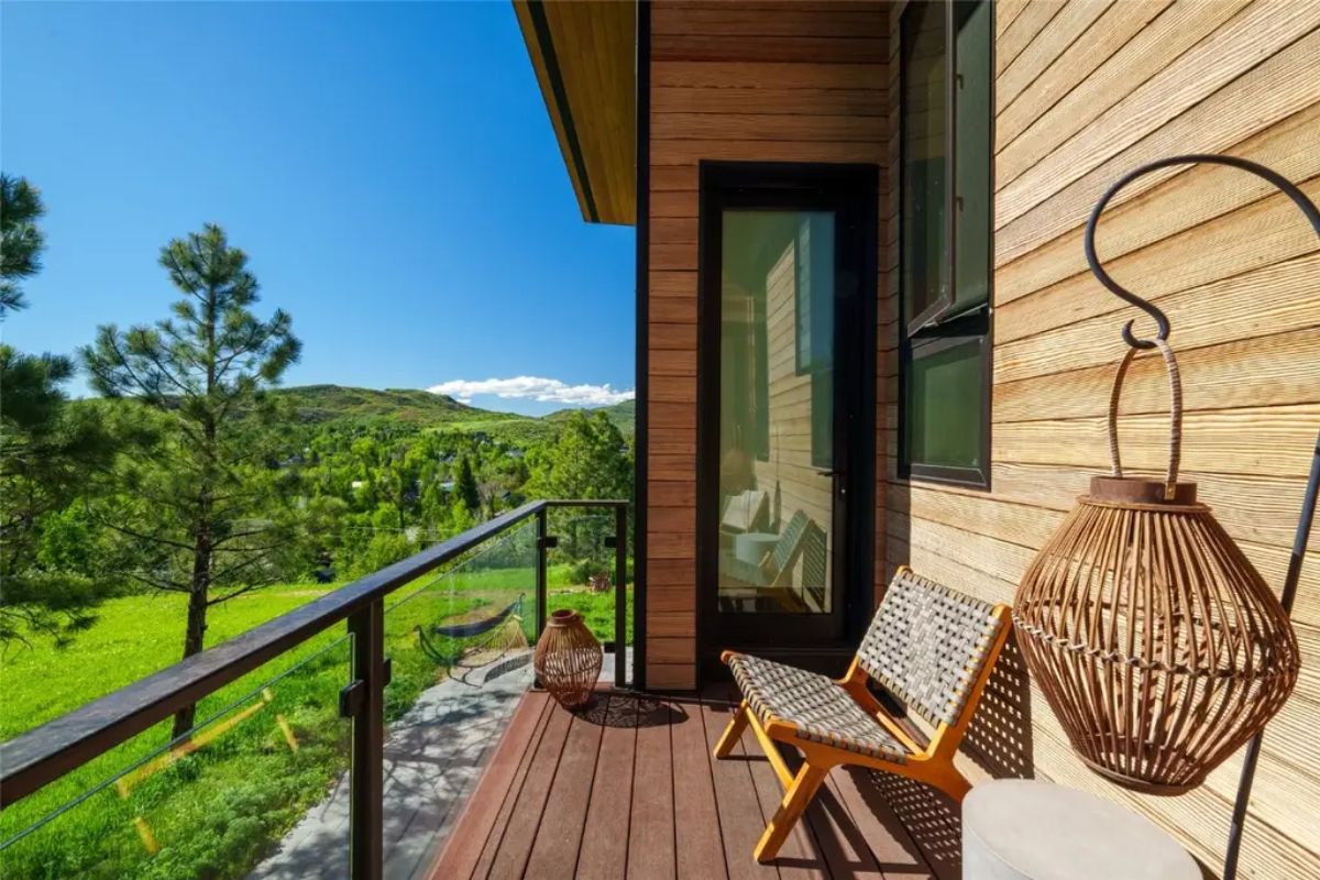 Small balcony with wooden siding, woven chairs, and glass railing overlooks a green landscape.
