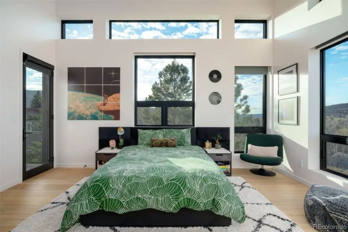 Bedroom with green bedspread and clerestory windows is framed by glass doors and views of surrounding forest.