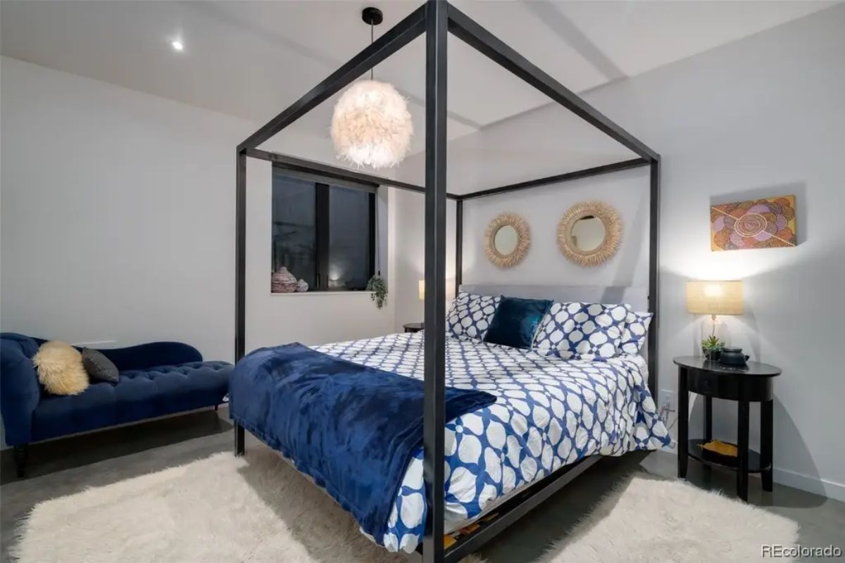 Four-poster bed with blue and white bedding sits on carpet next to a dark blue chaise lounge.