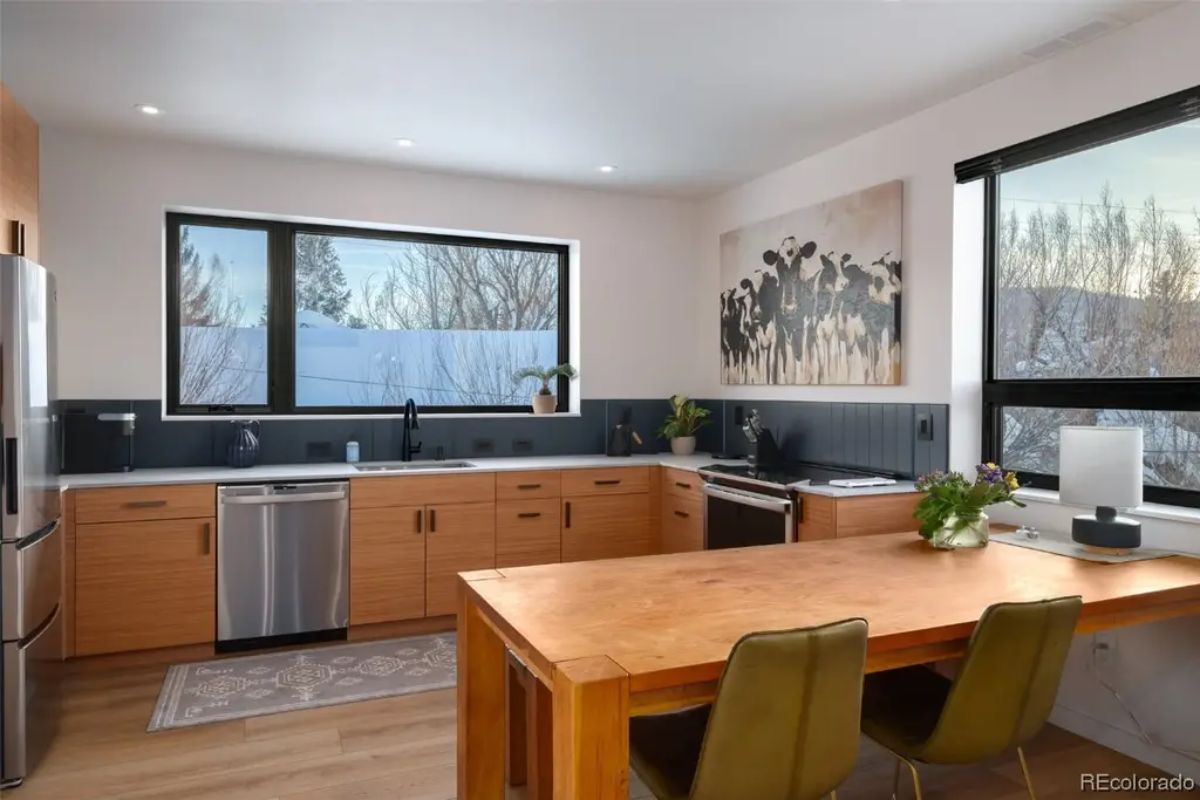 Second kitchen setup includes natural wood cabinetry, stainless steel appliances, and integrated dining table under large windows.