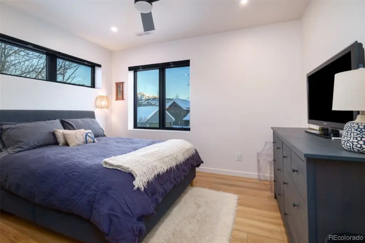 Bedroom with dark blue bedspread, wood flooring, and flat-screen TV looks out to snow-capped peaks through two windows.