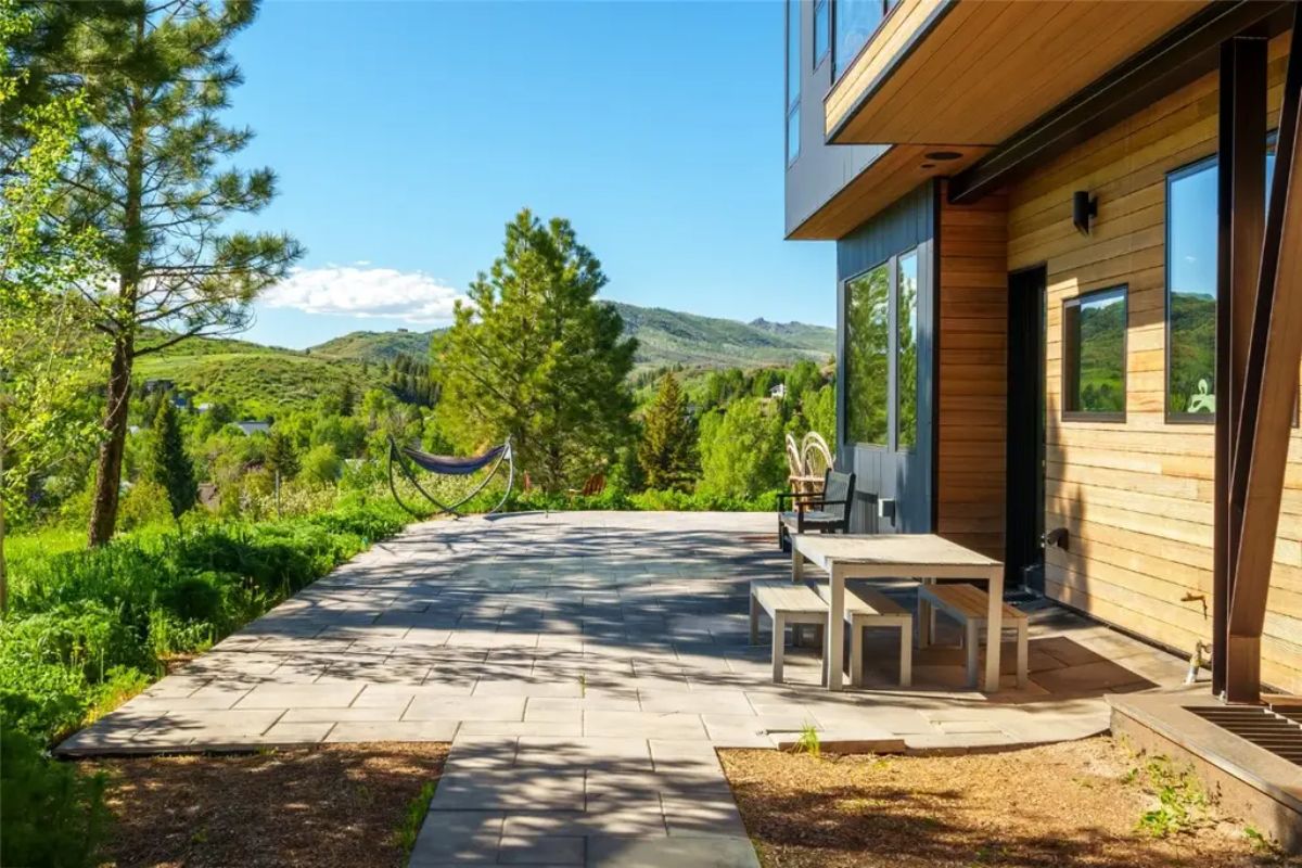 Rear stone patio with picnic table, chairs, and hammock set against wooded hillside.