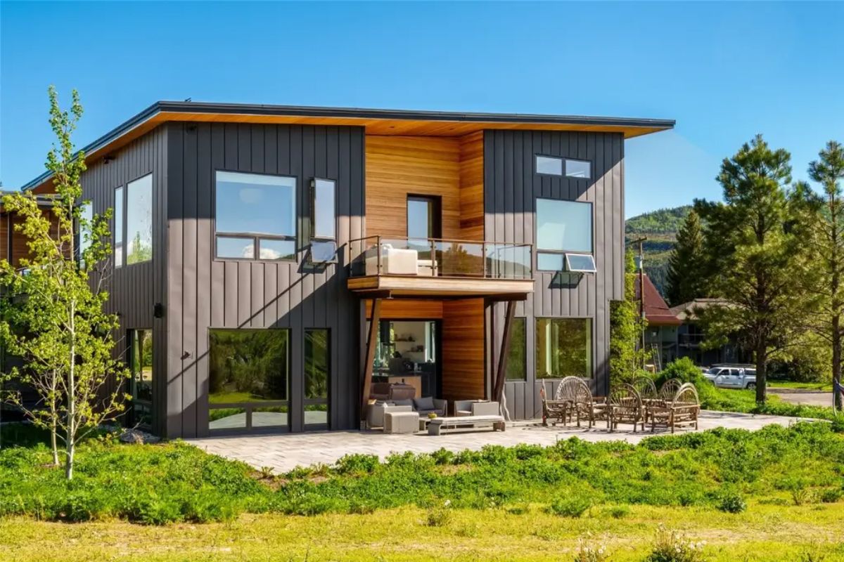 Two-story home with mixed metal and wood exterior has balcony, patio seating, and dining area.