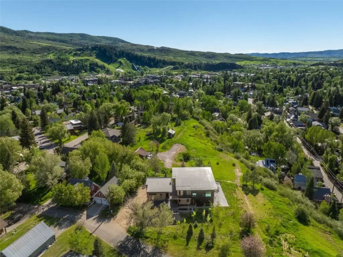 Elevated aerial view shows house centered in large green lot with nearby neighborhood and hills beyond.