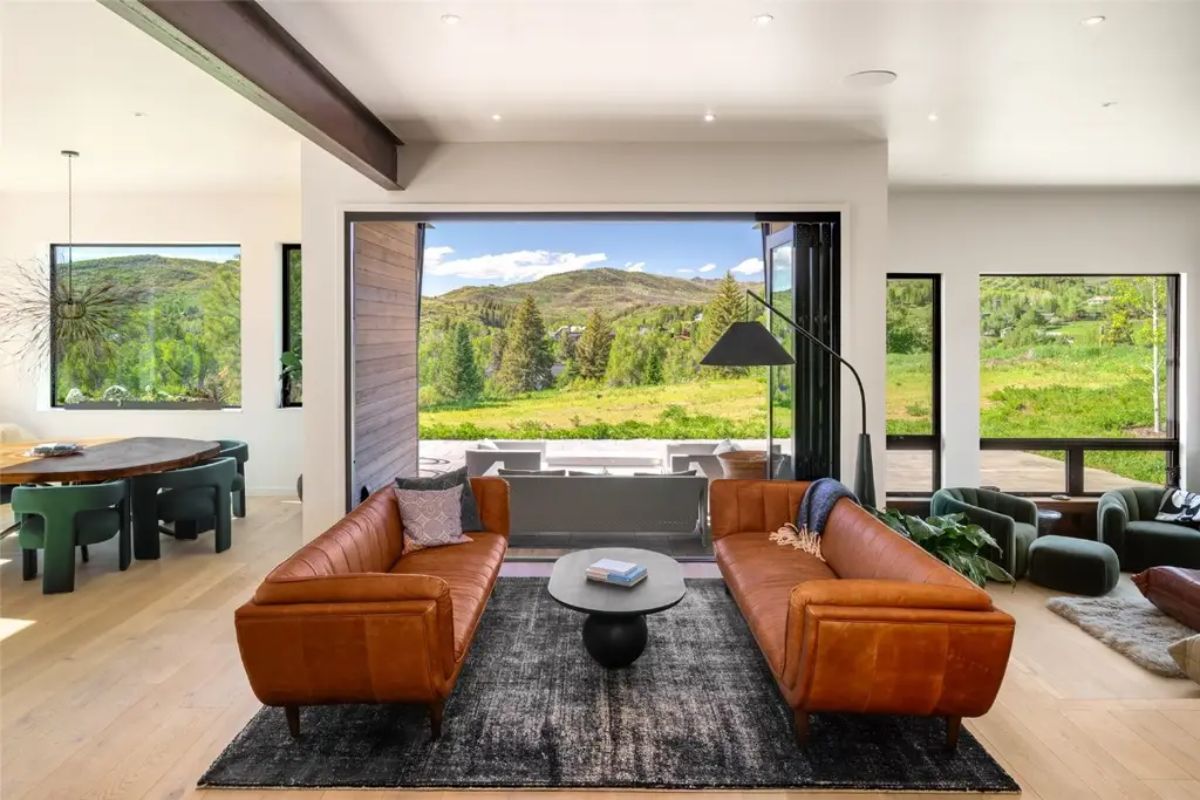 Living room with two brown leather sofas and large sliding window opens to a patio with mountain and forest views.