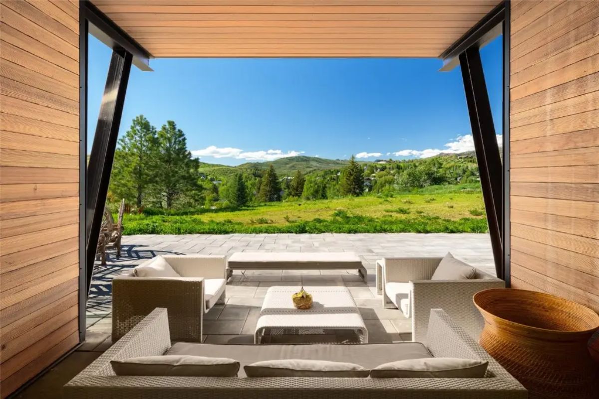 Covered patio with modern furniture faces grassy open space and tree-lined hills.