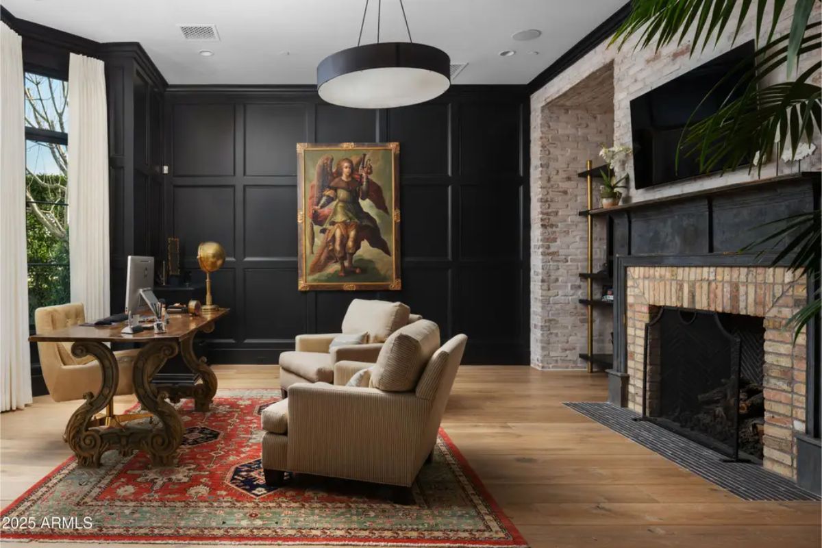 Home office with brick fireplace, dark paneled walls, and framed art above two armchairs.