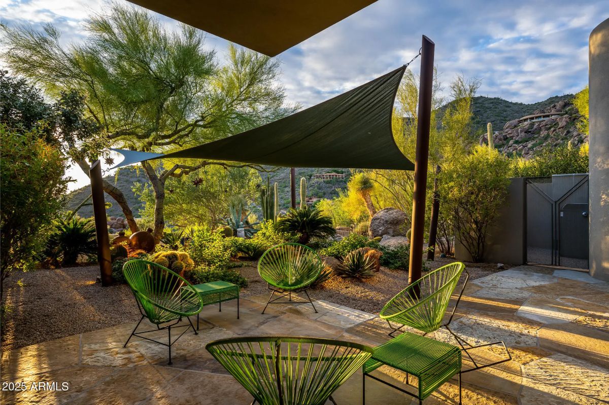 Outdoor sitting area with green chairs beneath triangular shade sails and surrounded by desert plants.