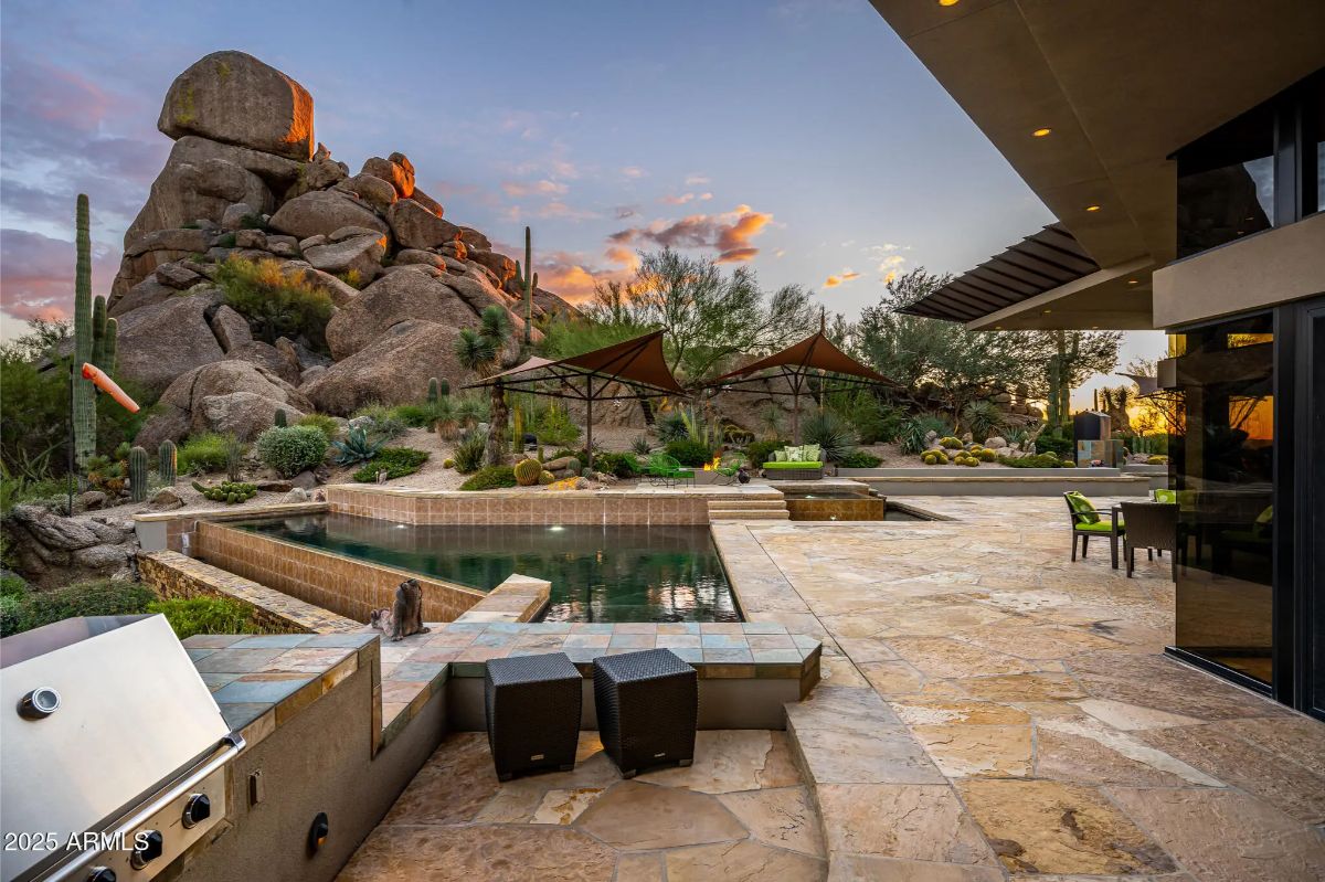 Stone patio with built-in grill, seating areas, and pool near large rock formations.