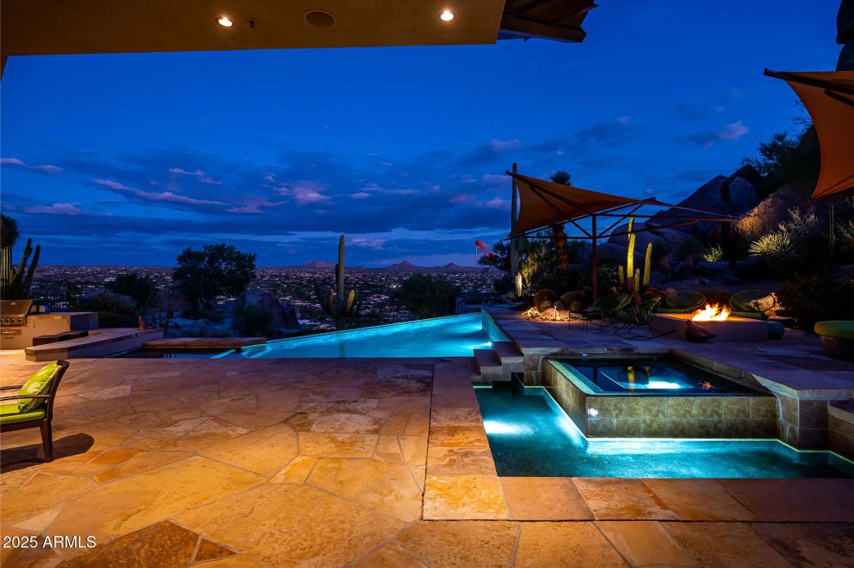 Evening poolside view with illuminated spa, fire pit, and distant city lights.
