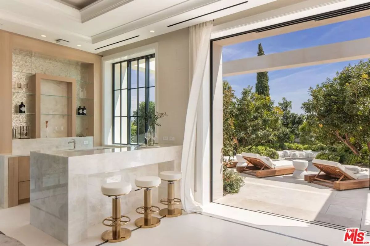 Indoor lounge bar with marble counter and barstools opens to patio with daybeds and garden view.
