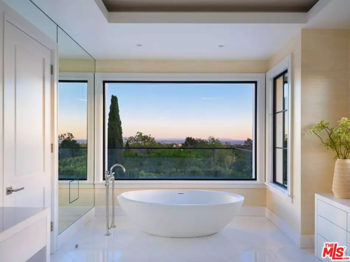 Bathroom with freestanding tub faces full-height picture window with treetop and skyline view.