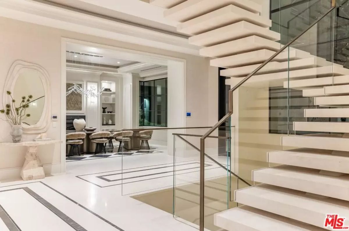 White staircase with glass railing rises beside dining area with light-toned chairs and geometric rug.