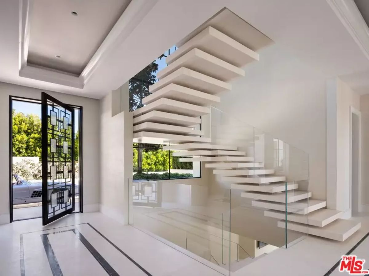 Floating staircase near pivot entry door is illuminated by floor-to-ceiling windows and glass railing.