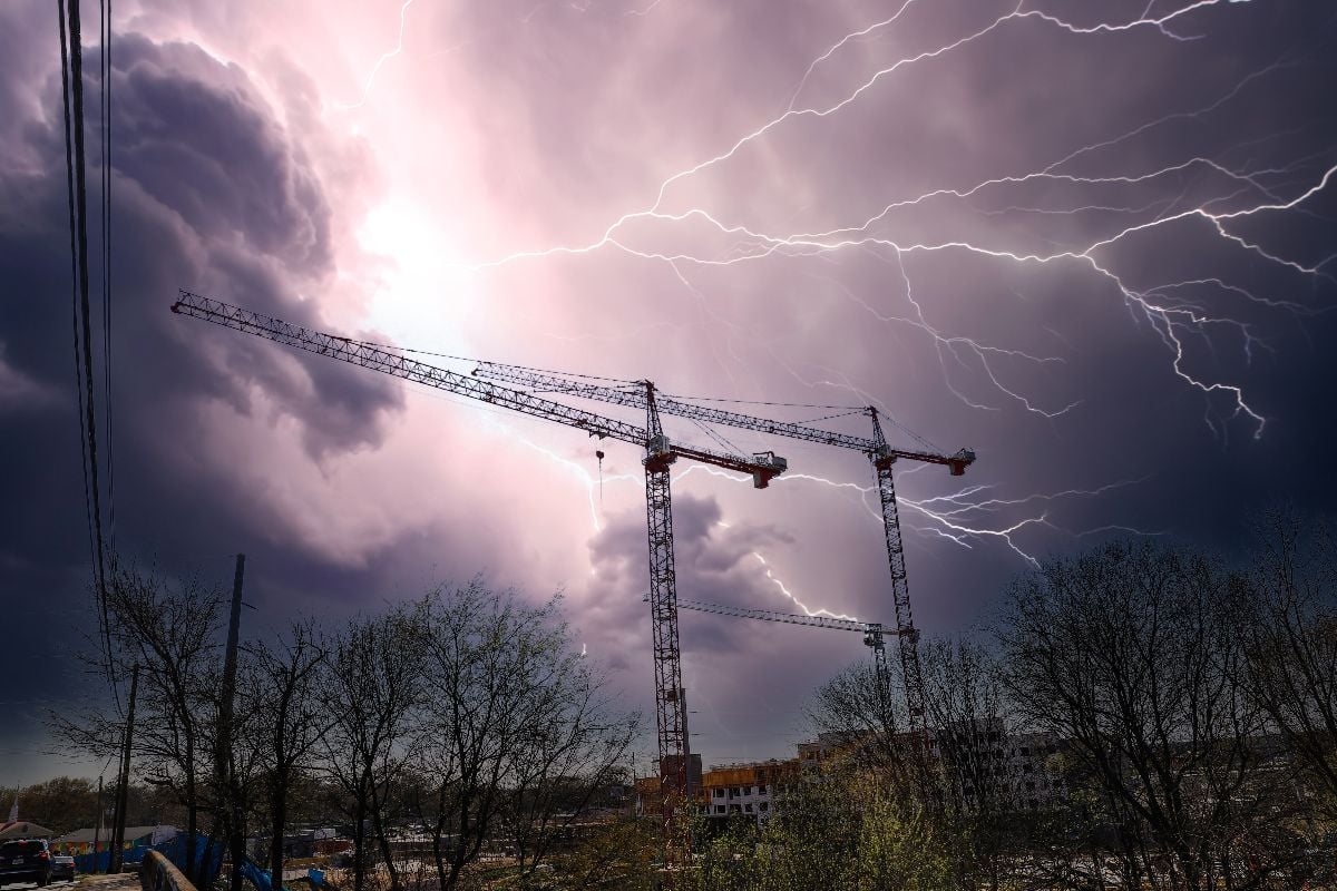 Tall black tower cranes at a construction site surrounded by lush green and bare winter trees and buildings.