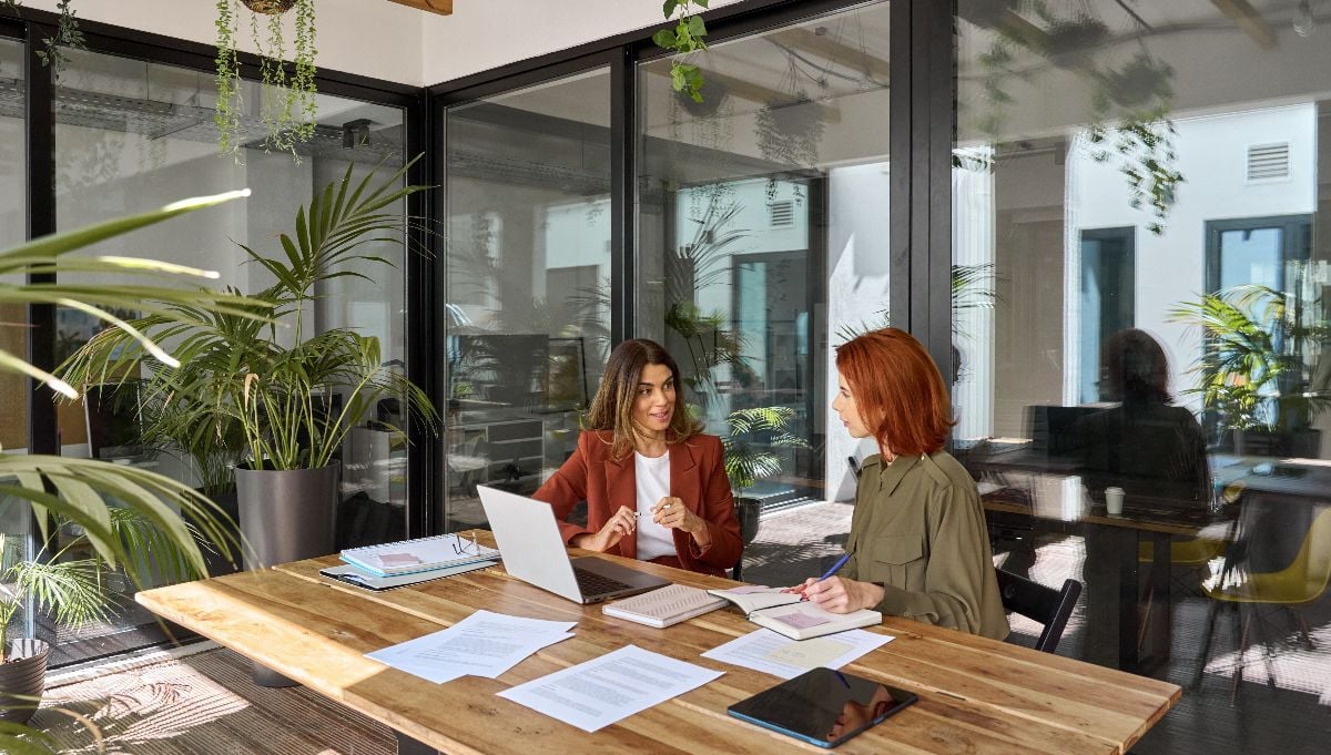 Two busy business women talking in creative green office sitting at desk.