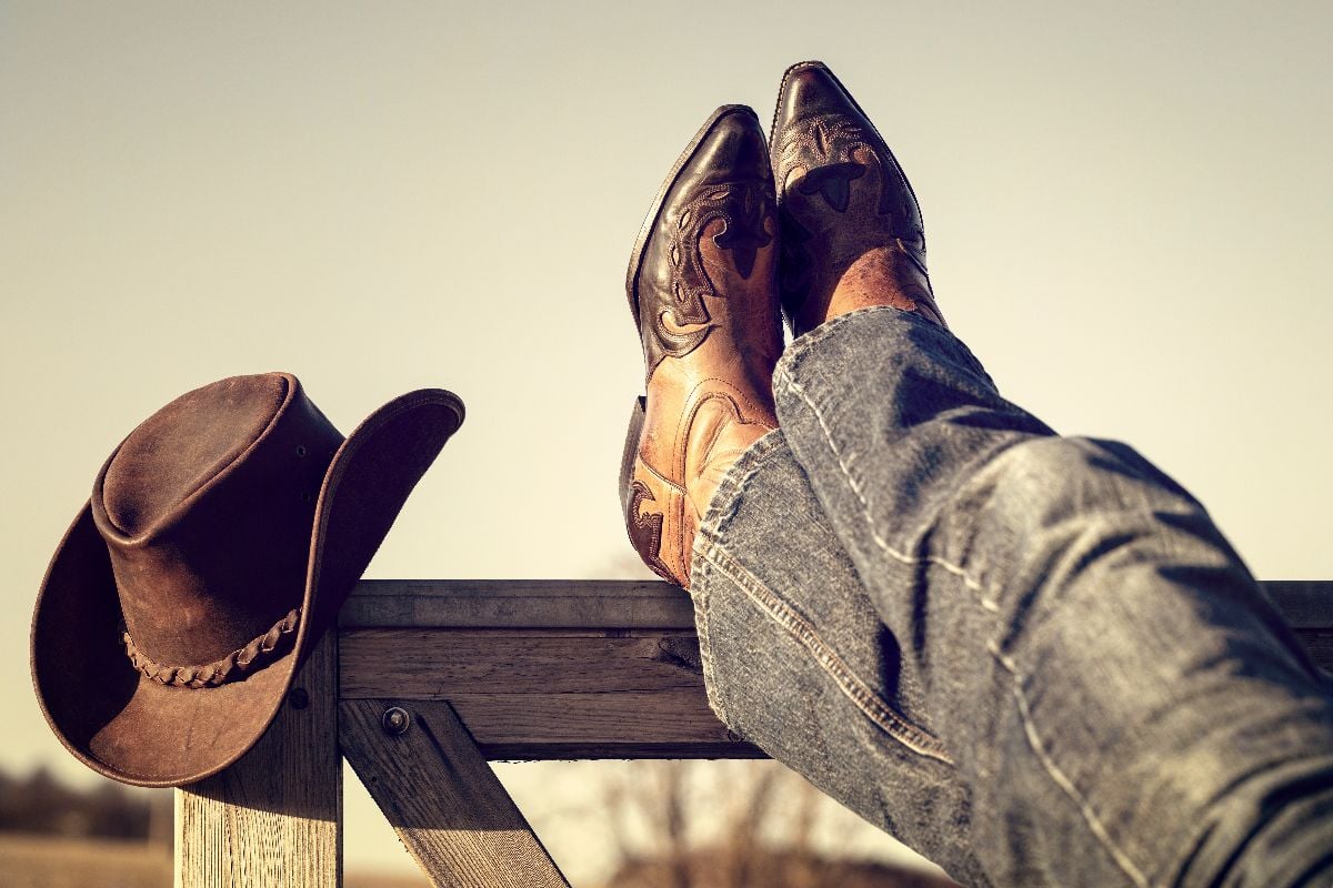 Cowboy boots and hat with feet up on stables gate at ranch.
