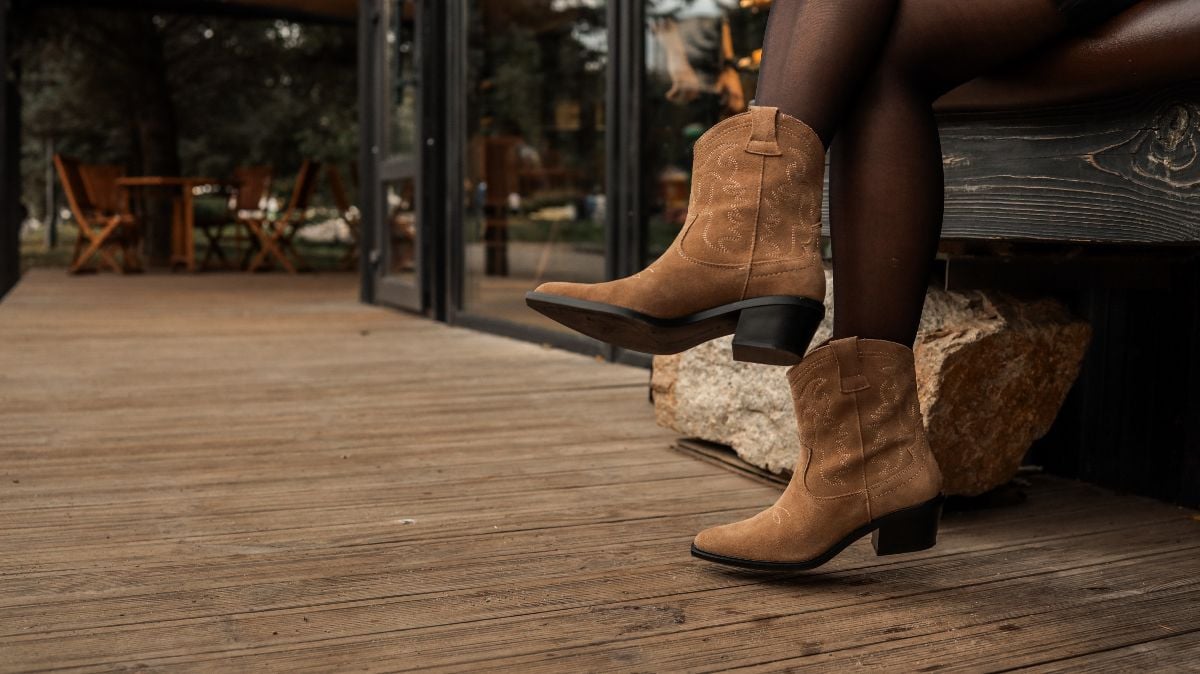 A woman sitting on a wooden bench, wearing a pair of brown suede cowboy boots.