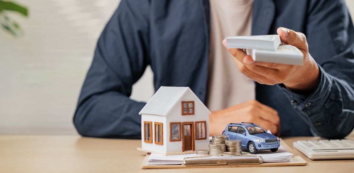 A man with cash bills, house, and car models on a table.