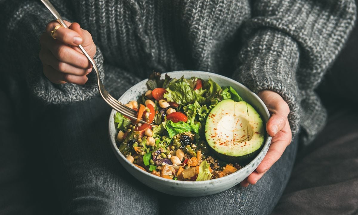 Woman in jeans and warm sweater holding bowl with fresh salad, avocado, grains, beans, and roasted vegetables.