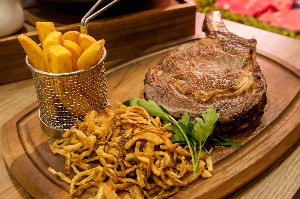 Large tomahawk steak, chips and tobacco onions, presented on a wooden chopping board and wire basket.