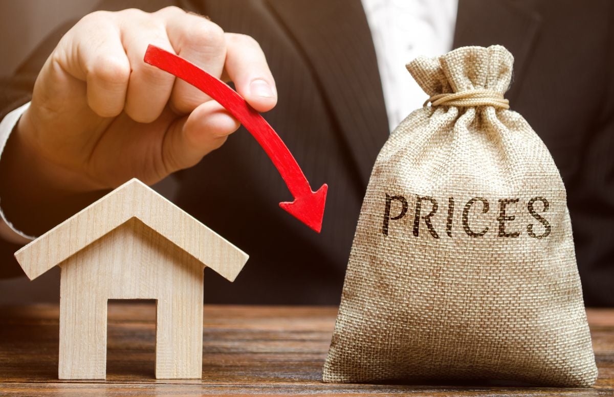 A bag with the word Prices, a miniature house and a down arrow in the hands of a businessman. 