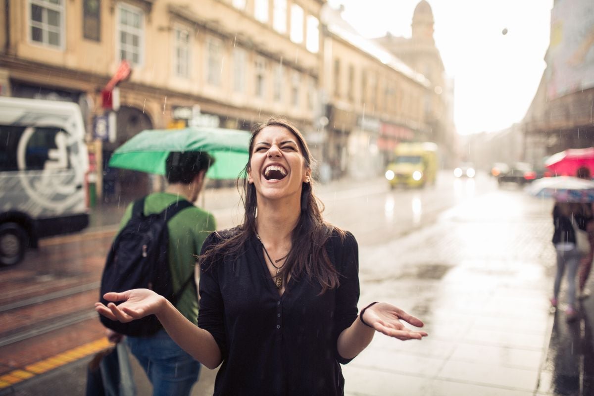 Woman standing on the street with her hands spread enjoying rain.