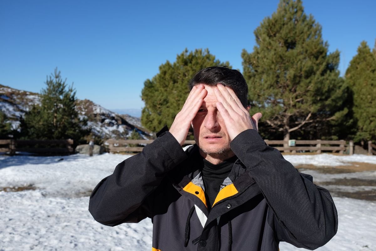  A man in a snowy landscape, wearing a black winter jacket, and applying sunscreen to his face.