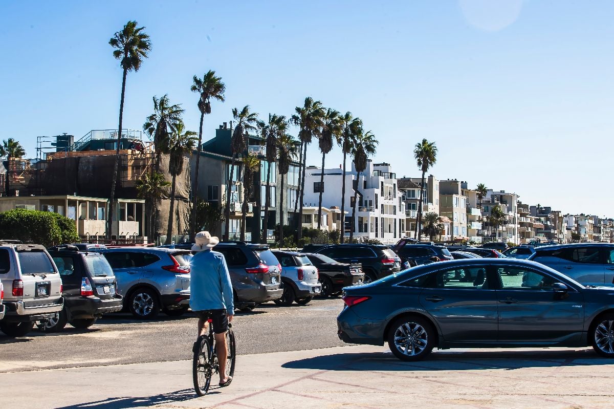 A person on a bike rides past a crowded beach parking lot with cars.